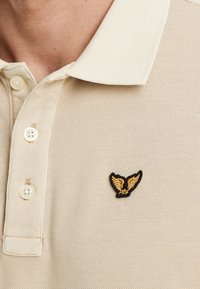 Beżowa koszulka polo o teksturowanej tkaninie, z kołnierzykiem zapinanym na trzy guziki oraz czarno-złotym logo w kształcie skrzydła na klatce piersiowej.