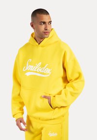 Felpa gialla in misto cotone, con un logo bianco, tasca frontale e polsini e orlo a coste. Abbinata a pantaloni jogger coordinati.