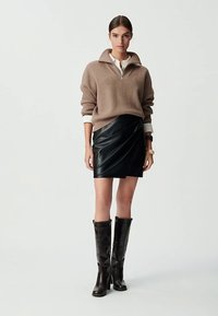 Suéter de punto beige con textura acanalada y cuello con cremallera, combinado con una falda negra de cuero envolvente. Botas negras de cuero hasta la rodilla completan el conjunto.