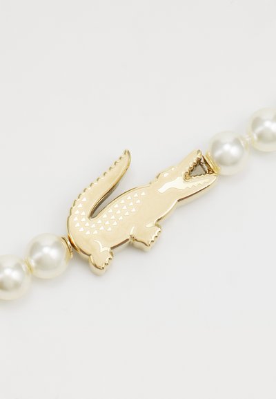 Clasp en forme de crocodile en or avec des détails texturés, associé à des perles de nacre blanches, créant une pièce de bijouterie distinctive.