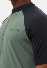 Zielono-granatowa koszulka rowerowa z zamkiem, wykonana z gładkiego materiału; ma zaokrąglony kołnierz i logo "Vaude" z przodu.