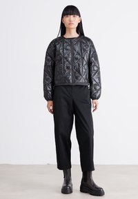 Vrouw met lang zwart haar, gekleed in een zwarte gewatteerde jas, zwarte cropped broek en zwarte chunky laarzen, staande tegen een effen witte achtergrond.