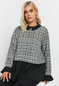 Maglione a righe bianche e nere con colletto rotondo nero, maniche lunghe e design a strati; il tessuto testurizzato crea interesse visivo.