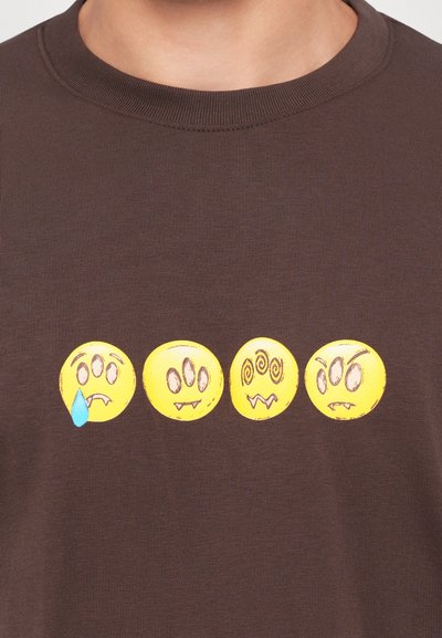 Štyri žlté emotikony s tromi očami na hnedom tričku, ktoré prejavujú smutnú tvár so slzou, neutrálnu tvár, zamotanu tvár a nahnevanú tvár.