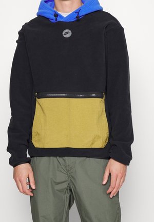 Personne portant un pull zippé noir et jaune avec une capuche bleue et un pantalon vert olive, debout sur fond blanc.