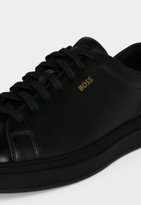 Baskets en cuir noir avec lacets noirs et logo doré "BOSS" sur le côté, montrant l’avant et le côté extérieur de la chaussure.