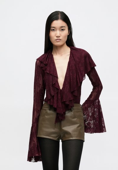 Bordowa koronkowa bluzka z falbanką przy dekolcie i rękawami dzwonkowymi, zestawiona z oliwkowozielonymi krótkimi spodniami z eko skóry oraz czarnymi rajstopami.