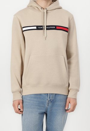 Sweat à capuche beige avec poche avant, bande horizontale noire, blanche et rouge sur la poitrine avec texte blanc, porté avec un jean bleu clair.
