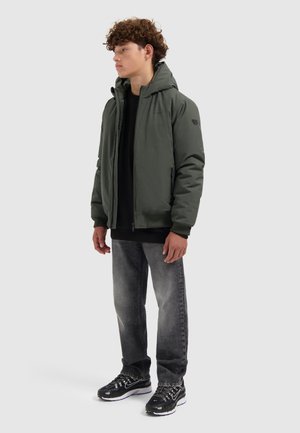 Jonge man in een groene hooded jacket, zwarte sweatshirt, vervaagde jeans en zwarte sneakers die tegen een effen lichte achtergrond staat.