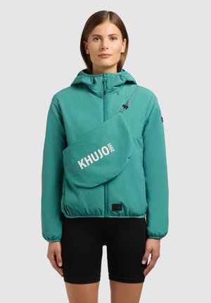 Kvinde iført teal lynlåsjakke med hætte og matchende crossbody taske mærket "KHUJO", kombineret med sorte cykelshorts, stående mod hvid baggrund.