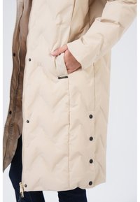Beige gewatteerde jas met een rits en drukknoopjes, voorzien van een zijzak en een soepele textuur. Geen zichtbare patronen of accenten.