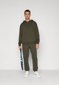 Sweatshirt verde-oliva com um bolso frontal, combinado com calças de treino correspondentes. Ténis brancos. A segurar uma raquete de ténis azul e preta.