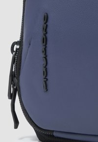 Bustina in pelle blu con superficie strutturata, dotata di cerniera nera e logo in rilievo. Include cuciture nere lungo i bordi.