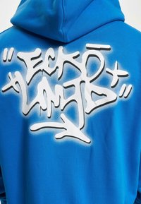 Un hoodie bleu en coton avec un design blanc de style graffiti au dos, présentant des formes abstraites et des éléments de texte en accents noirs.