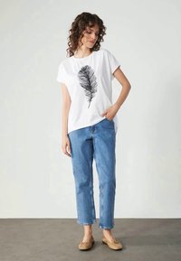Hvit kortermet t-skjorte med svart fjærtrykk, kombinert med lyseblå rette jeans og beige leiligheter. Enkel bakgrunn.