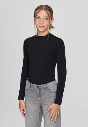 Maglietta nera a maniche lunghe con collo a imbuto realizzata in tessuto morbido, abbinata a jeans grigio chiaro con dettagli di cuciture minimali.