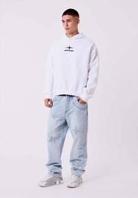 Sudadera blanca con el logo "PXA" en negro, jeans de un tono azul claro con costuras en forma de estrella, y zapatillas grises. Ajuste relajado en general.