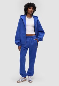 Niebieska bluza z kapturem na zamek, duże kieszenie i luźny krój. W zestawie z niebieskimi dresami i srebrnymi sneakersami. Gładka tekstura.