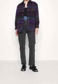 Camisa de franela de cuadros en tonos púrpura, verde y marrón, con botones al frente, combinada con jeans rectos negros y zapatos negros brillantes.