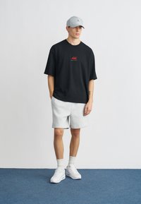 Μαύρο φαρδύ T-shirt με κόκκινο σχέδιο, ανοιχτόγκρι σορτς, λευκά αθλητικά παπούτσια και γκρι καπέλο. Φόντο ουδέτερο.