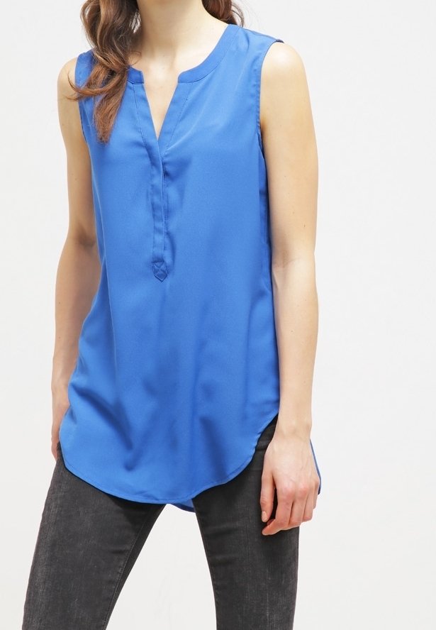 Femme portant un haut tunique bleu sans manches avec un col en V et un pantalon noir, debout devant un fond blanc uni.