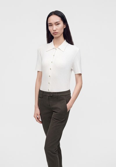 Femme aux longs cheveux foncés portant un haut blanc à manches courtes boutonné et un pantalon sombre, debout avec une main dans la poche devant un fond uni.