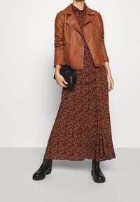Giubbotto da motociclista in pelle marrone sopra un vestito maxi nero con fiori rossi e rosa. Una pochette nera e stivaletti completano il look.