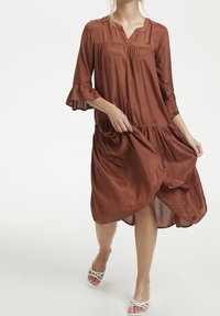 Brun, flydende kjole med V-udskæring og trekvart lange klokkeærmer. Har et lagdelt design, blød tekstur og en afslappet pasform.
