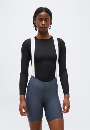 Femme portant un haut noir à manches longues avec des bretelles blanches et un cuissard de cyclisme gris taille haute, debout devant un fond uni.
