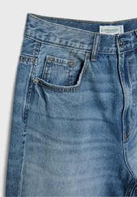 Jean en denim bleu gros plan montrant les poches avant, les passants de ceinture, le bouton en métal et l'étiquette de la marque Stradivarius sur la ceinture.