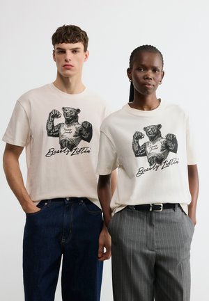 Young man and woman wearing white graphic tees with muscular bear illustration and "Bearly Lifting" text, standing against plain background.