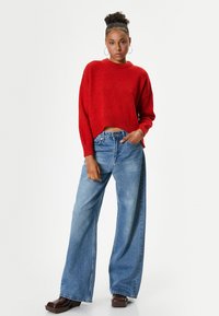Rode cropped sweater met een ronde hals, gecombineerd met wijde blauwe denim jeans. Model draagt bruine platformschoenen en oorringen.