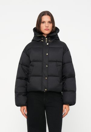 Giacca a vento nera con cappuccio, caratterizzata da un design quadrato, bottoni a scatto dorati e texture trapuntata. Indossata con pantaloni scuri.