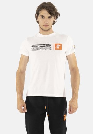 Jeune homme portant un t-shirt blanc avec le texte répété "We are combat sports" et un pantalon noir avec des détails orange.