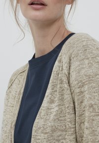 Cardigan en tricot beige avec un motif texturé, superposé sur une chemise bleu marine. Gros plan des détails du col et de l'épaule.