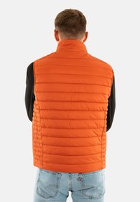 Oranje geïsoleerde vest met een gestikt ontwerp, voorzien van verticale stiksels en een hoge kraag, gedragen over een zwart shirt met lange mouwen.