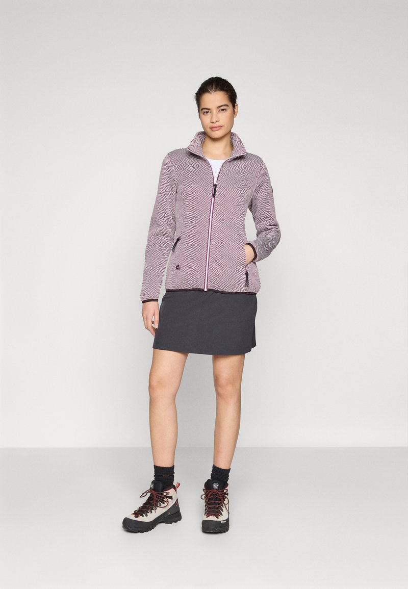 Giacca in fleece con zip, caratterizzata da un motivo geometrici in rosa e bianco, abbinata a una gonna grigia e a scarponi da trekking. Dettagli in tessuto testurizzato e colletto visibili.
