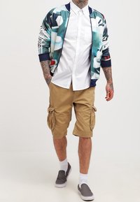 Veste bomber imprimée avec des accents verts, bleus et rouges sur une chemise blanche à boutons, associée à un short cargo beige et des chaussures gris slip-on.
