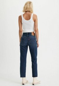 Jeans de mezclilla azul oscuro con corte recto, que presentan bolsillos traseros y un parche de cuero con el logo. Combinados con una camiseta sin mangas blanca y zapatillas beigas.