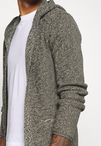 Szary dzianinowy kardigan z kapturem, o fakturowanym splocie, z otwartym przodem i podwiniętymi rękawami. Noszony na białej koszulce.