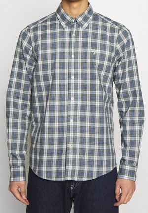 Mężczyzna ubrany w zieloną, białą i niebieską kratowaną koszulę na guziki z długimi rękawami oraz ciemne spodnie, stojący na jednolitym tle.