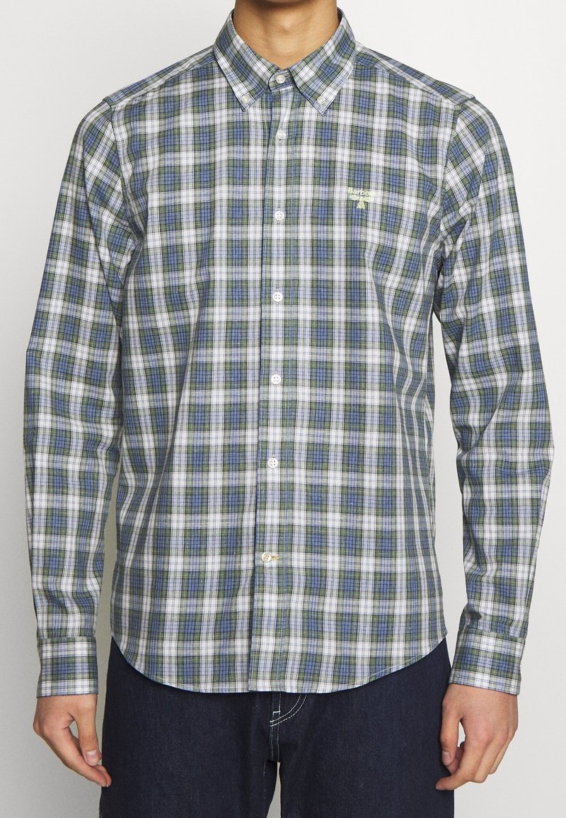 Homme portant une chemise à carreaux verts, blancs et bleus à manches longues et un pantalon foncé, se tenant devant un fond uni.