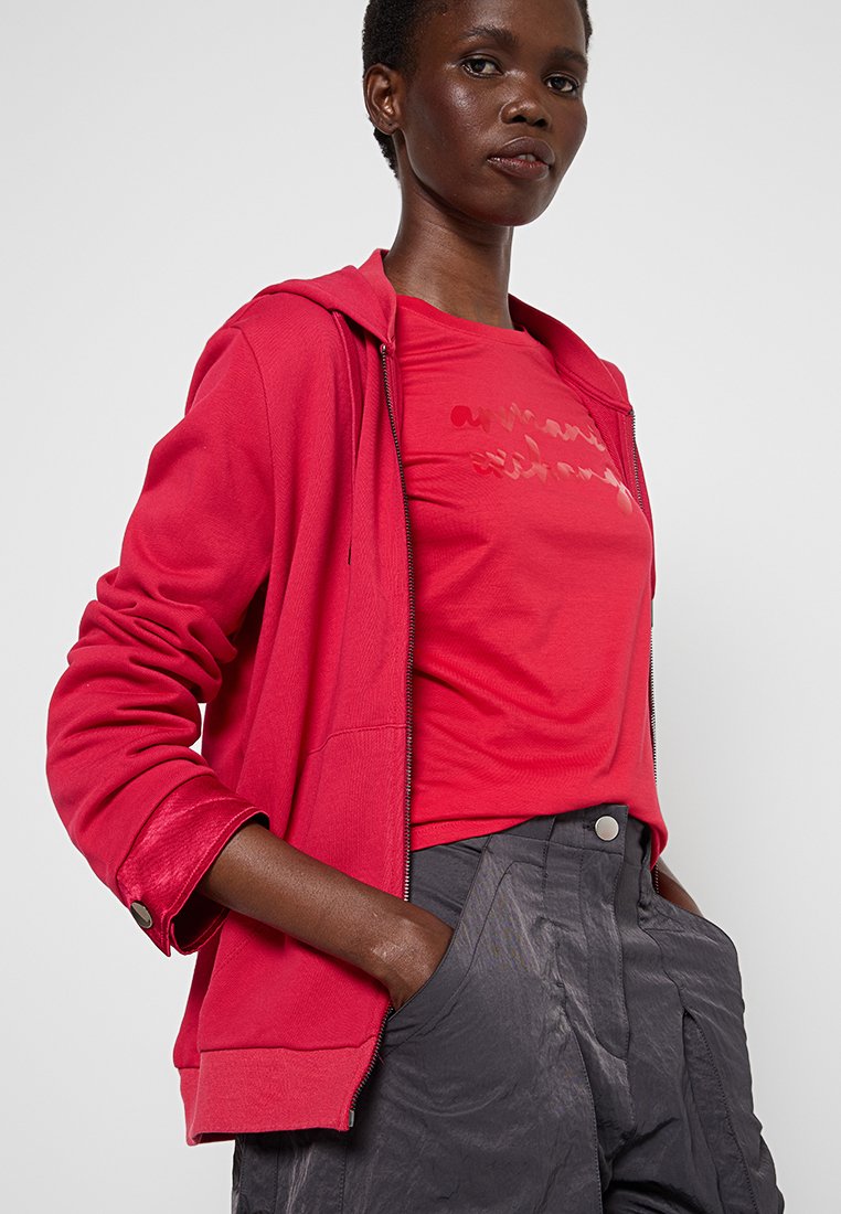 Donna che indossa una felpa rossa con zip sopra una maglietta rossa e pantaloni grigio scuro, con le mani nelle tasche, su uno sfondo chiaro e neutro.