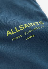 Niebieska tkanina o prążkowanej strukturze, z żółtym logotypem zawierającym napisy "ALLSAINTS" i "LONDYN" wydrukowane dużą czcionką.