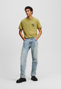 Camiseta verde oliva con un logo negro, combinada con jeans de mezclilla azul claro. El modelo lleva zapatos negros de suela gruesa, con las manos en los bolsillos.