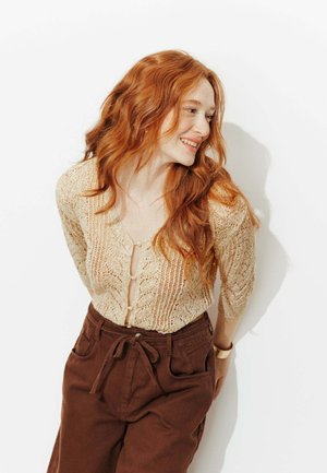 Femme rousse souriante appuyée contre un mur blanc, portant un cardigan en maille beige et un pantalon marron taille haute avec une ceinture nouée.