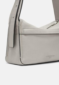 Jasnoszara skórzana torba na ramię o gładkiej fakturze, z zamkiem błyskawicznym i czarnymi akcentami. Logo marki wytłoczone z przodu.