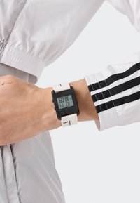 Digital klocka med en fyrkantig svart urtavla, vit silikonrem, med en stor LCD-skärm som visar tiden och subtila logotyper på remmen.