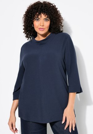 Haut en tricot bleu marine avec un col montant, des manches trois-quarts, et une texture côtelée, offrant une coupe décontractée et des fentes sur les côtés à l'ourlet.