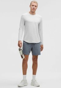 Jasnoszara koszula z długim rękawem o subtelnej fakturze w paski, w połączeniu z niebieskimi szortami. Trzyma przezroczystą butelkę na wodę. Białe buty sportowe.